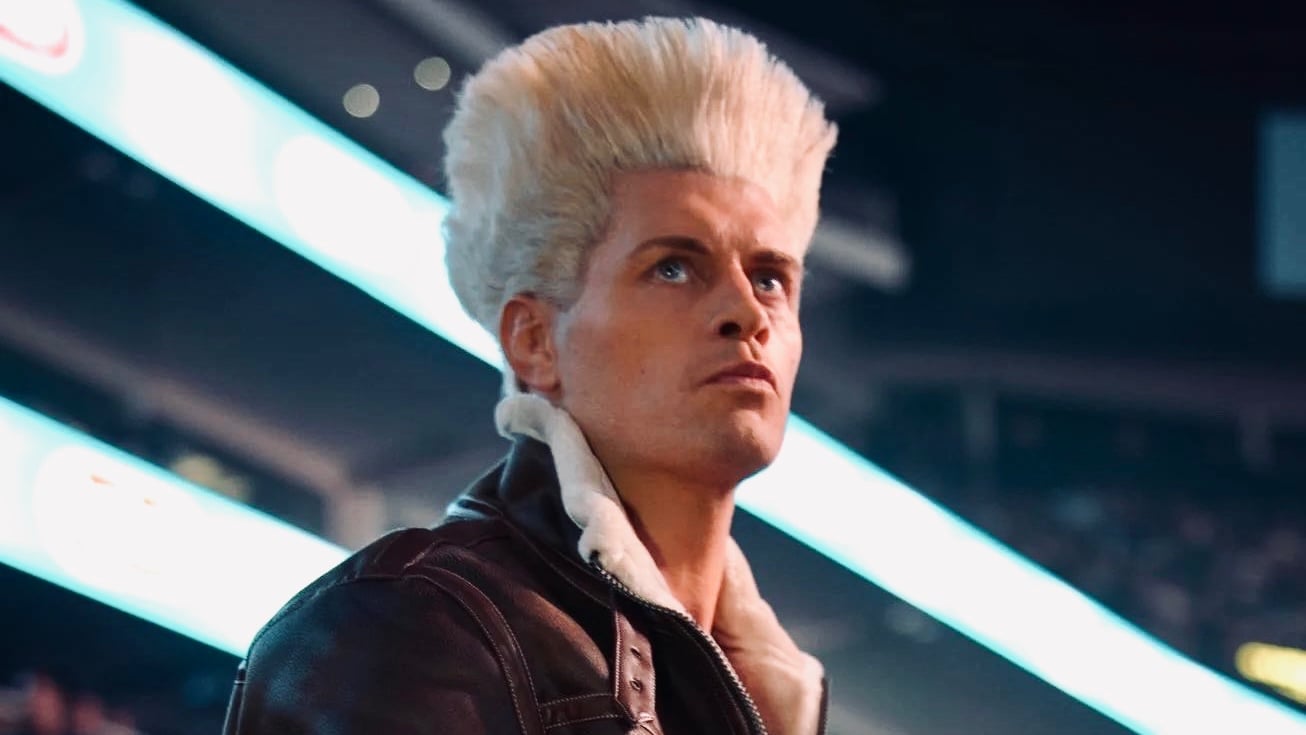Cody Rhodes dressed as Guile from Street Fighter, with a blond flat-top and leather bomber jacket, in an arena