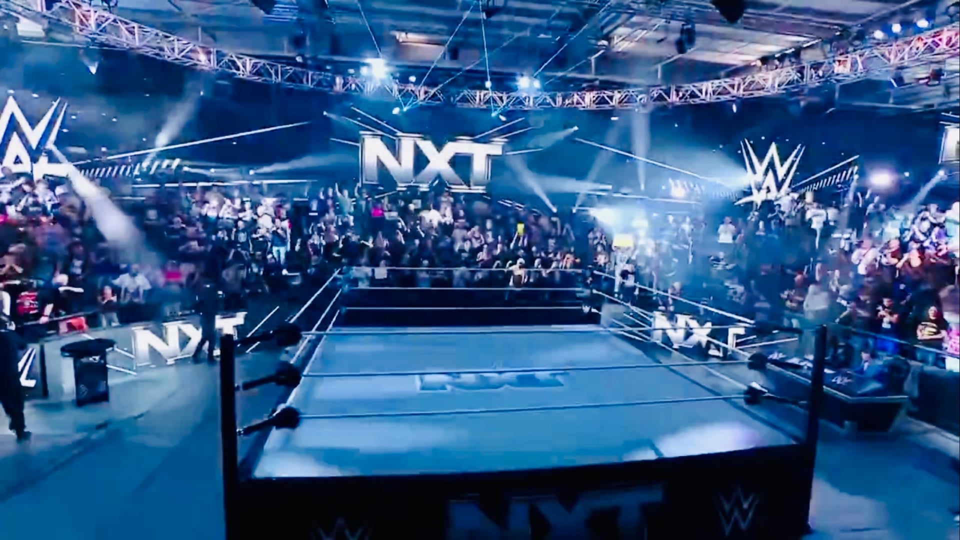 The NXT ring under bright stage lights with a packed crowd and NXT logos in the WWE Performance Center